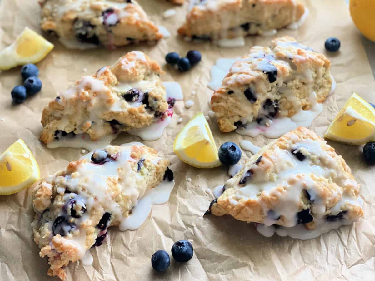 Six blueberry scones on brown paper with fresh blueberries and lemon wedges.