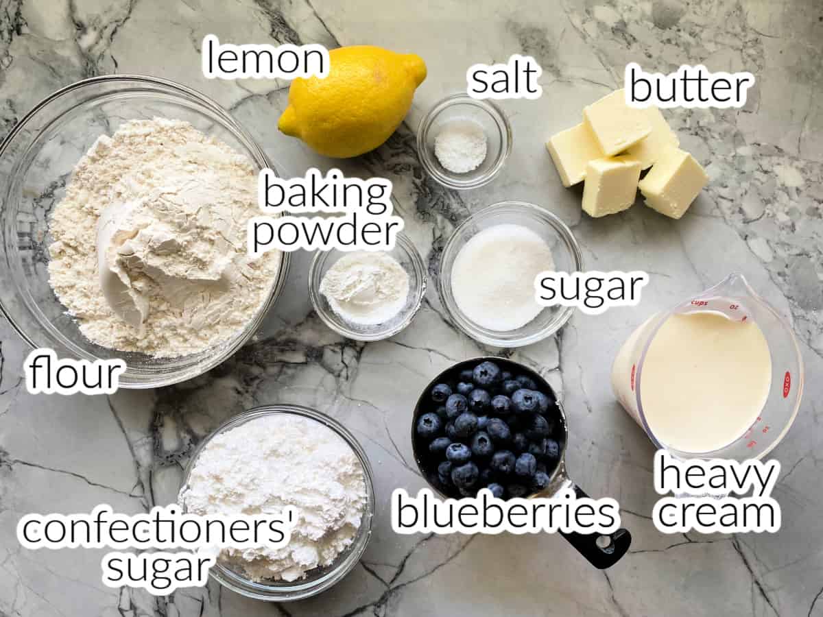 Ingredients on counter: flour, lemon, baking powder, confectioners' sugar, bluberries, salt, sugar, heavy cream, butter.