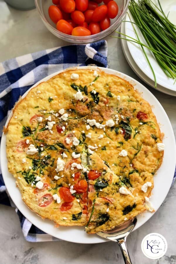 Egg fritatta with a slice being held by a silver serving utensil on a white plate with logo in the right corner.