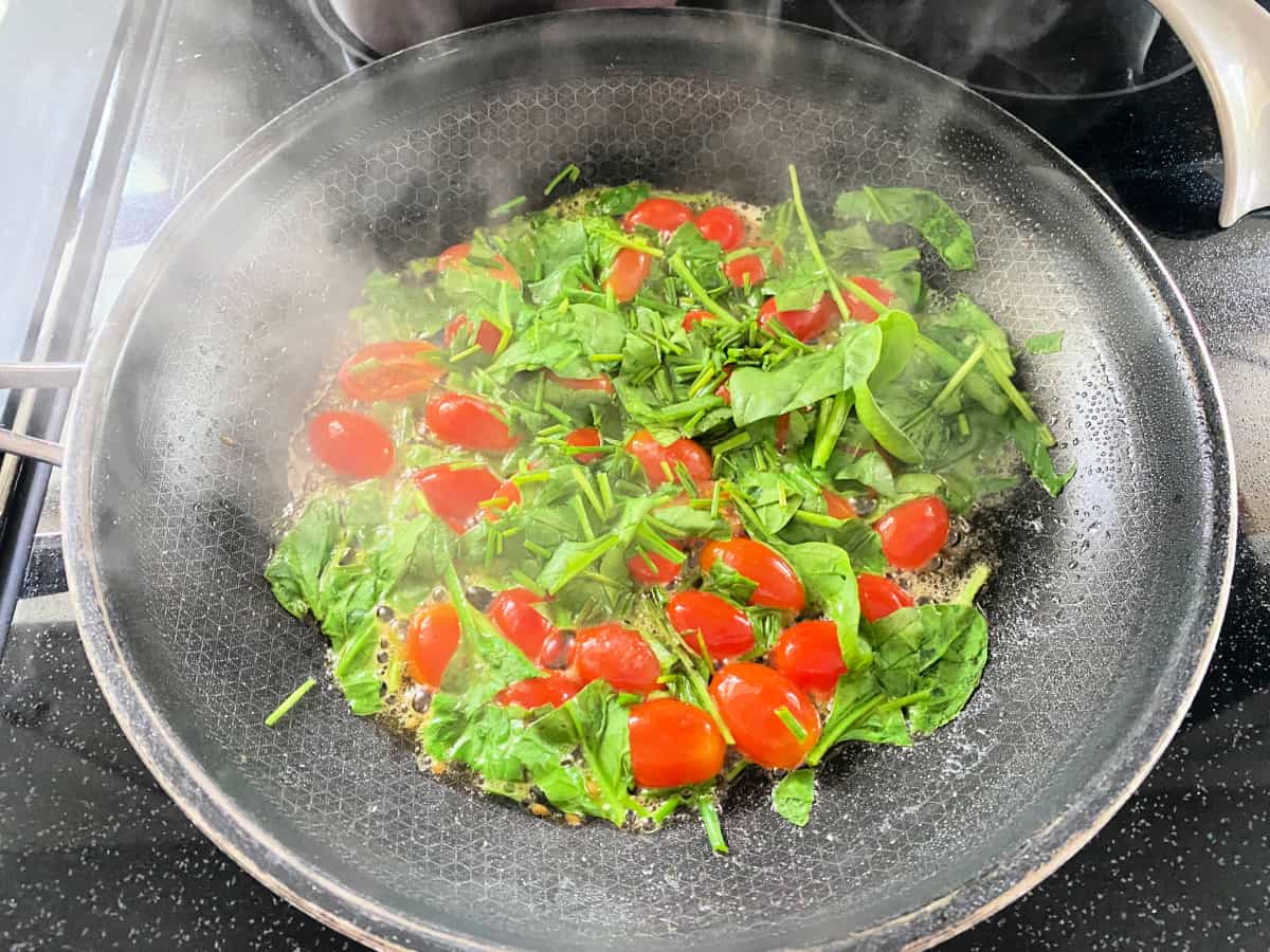 Black frying pan with steam coming from spinach and tomatoes.