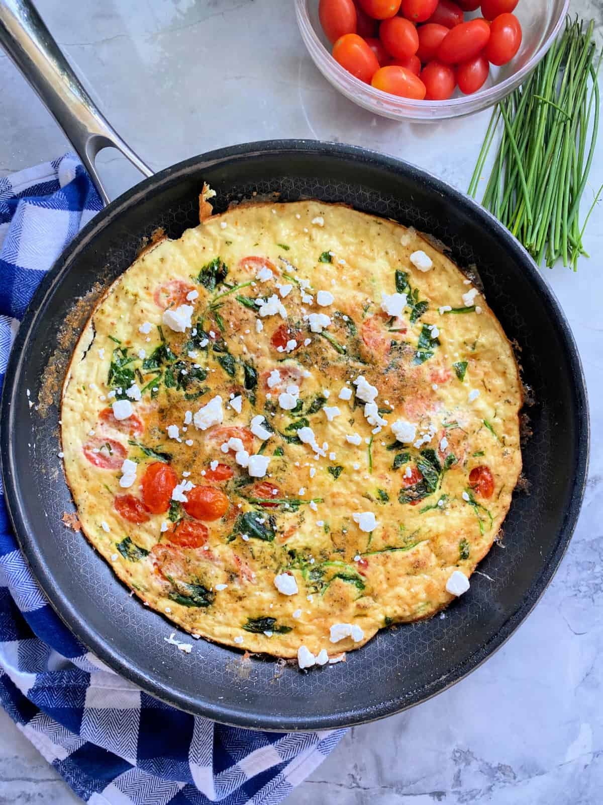 Black frying pan on a blue and white checkered cloth with an egg fritatta in the pan.