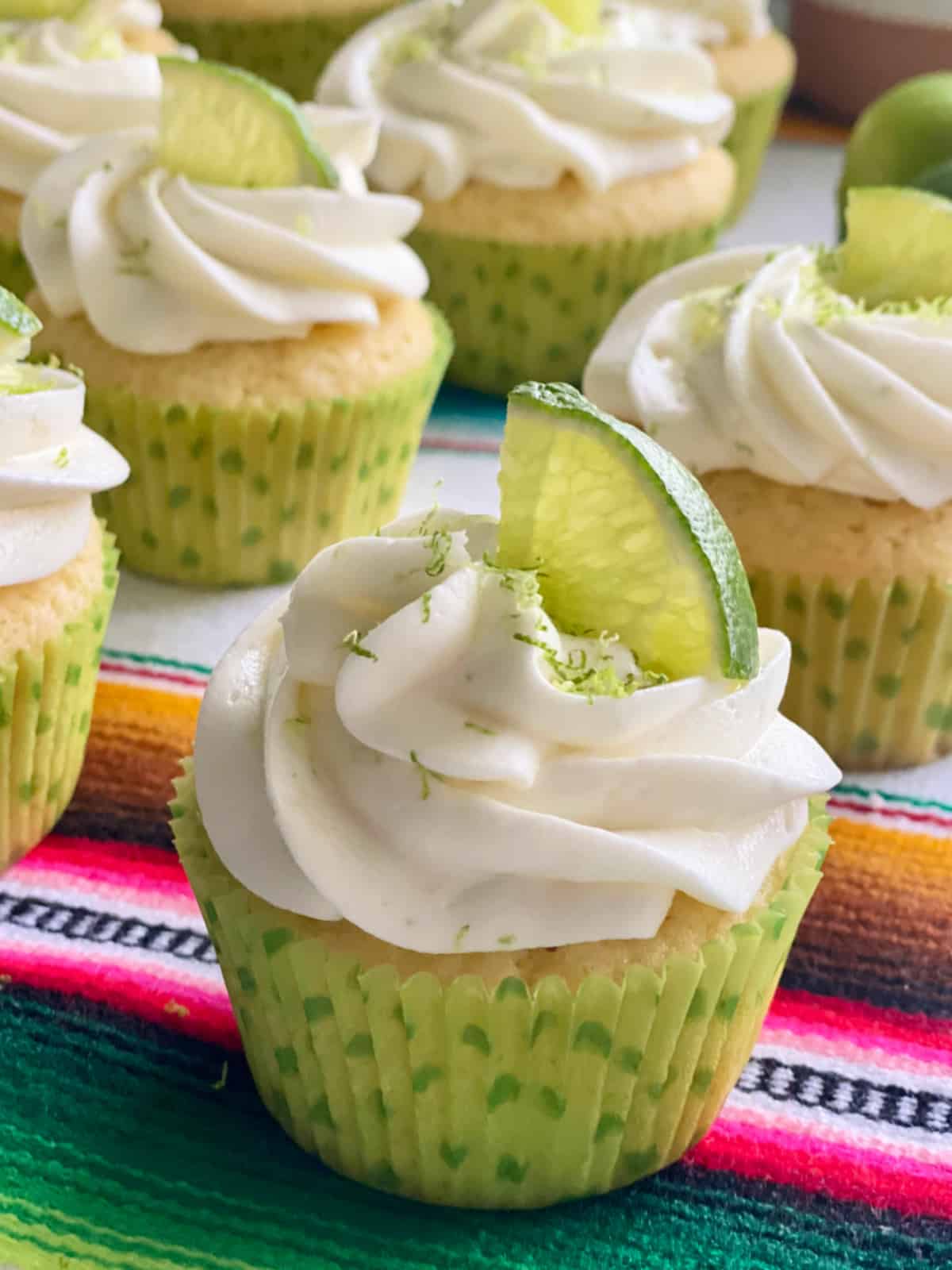 5 white cupcakes close up with white frosting and lime zest and lime on top in a lime green polka dot wrapper.