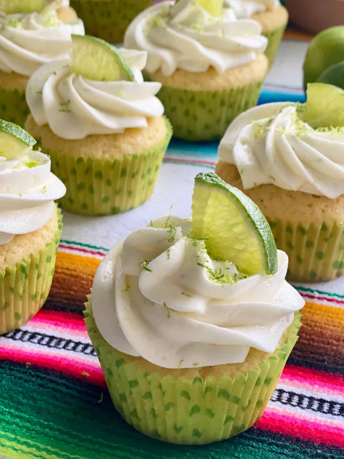 Seven white cupcakes with white frosting and lime zest and lime wedge on top in a green polka dot wrapper.