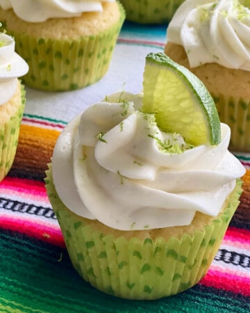 Four white cupcakes with white frosting and lime zest and lime on top in a green polka dot wrapper.