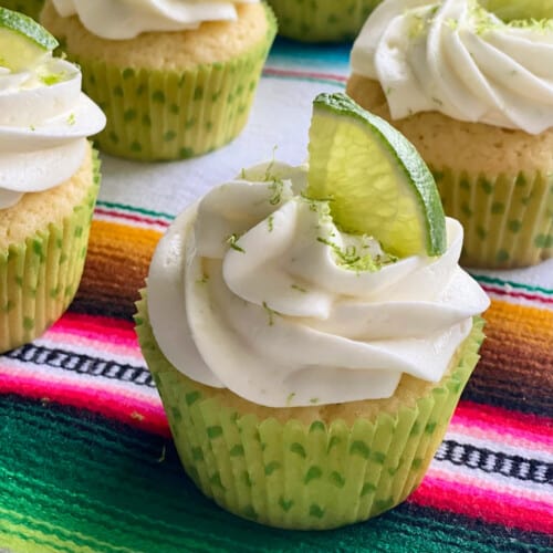 Four white cupcakes with white frosting and lime zest and lime on top in a green polka dot wrapper.