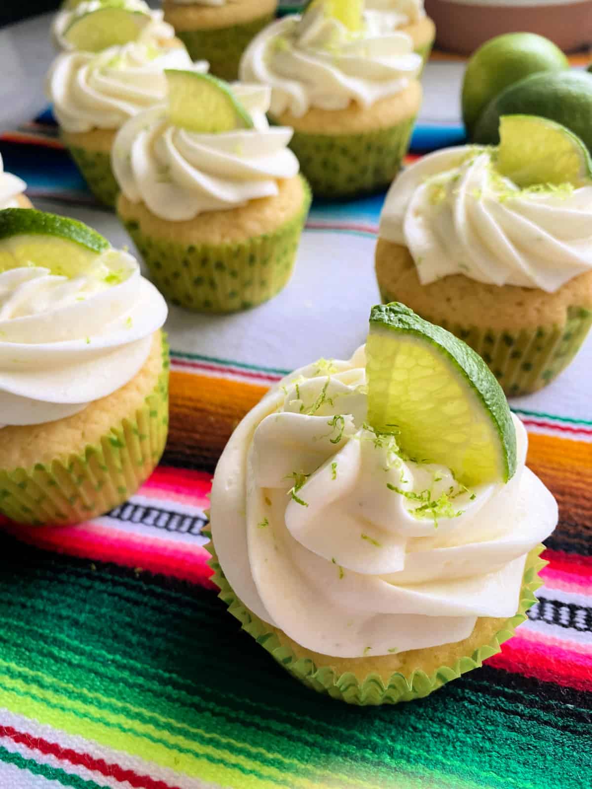 Eight white cupcakes with white frosting and lime zest and lime on top in a green polka dot wrapper.