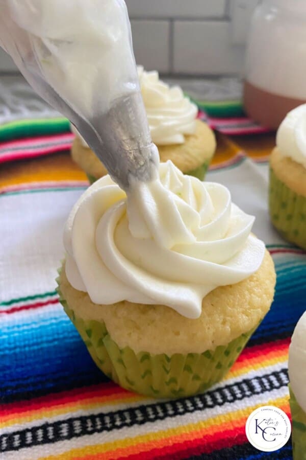 Frosting bag piping white frosting on to a cupcake with logo on bottom right corner.