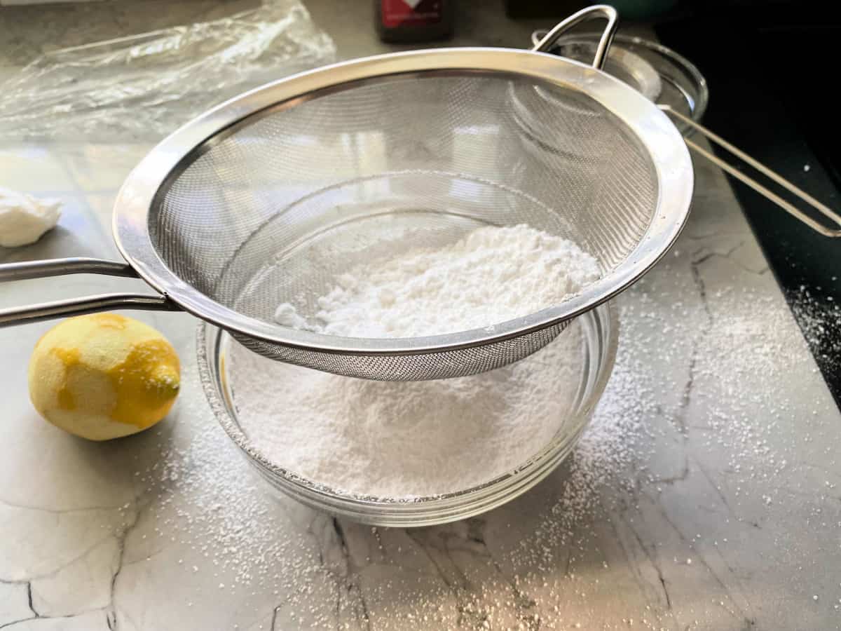 Mesh sifter with confectioners' sugar with glass bowl filled with sugar on a countertop.