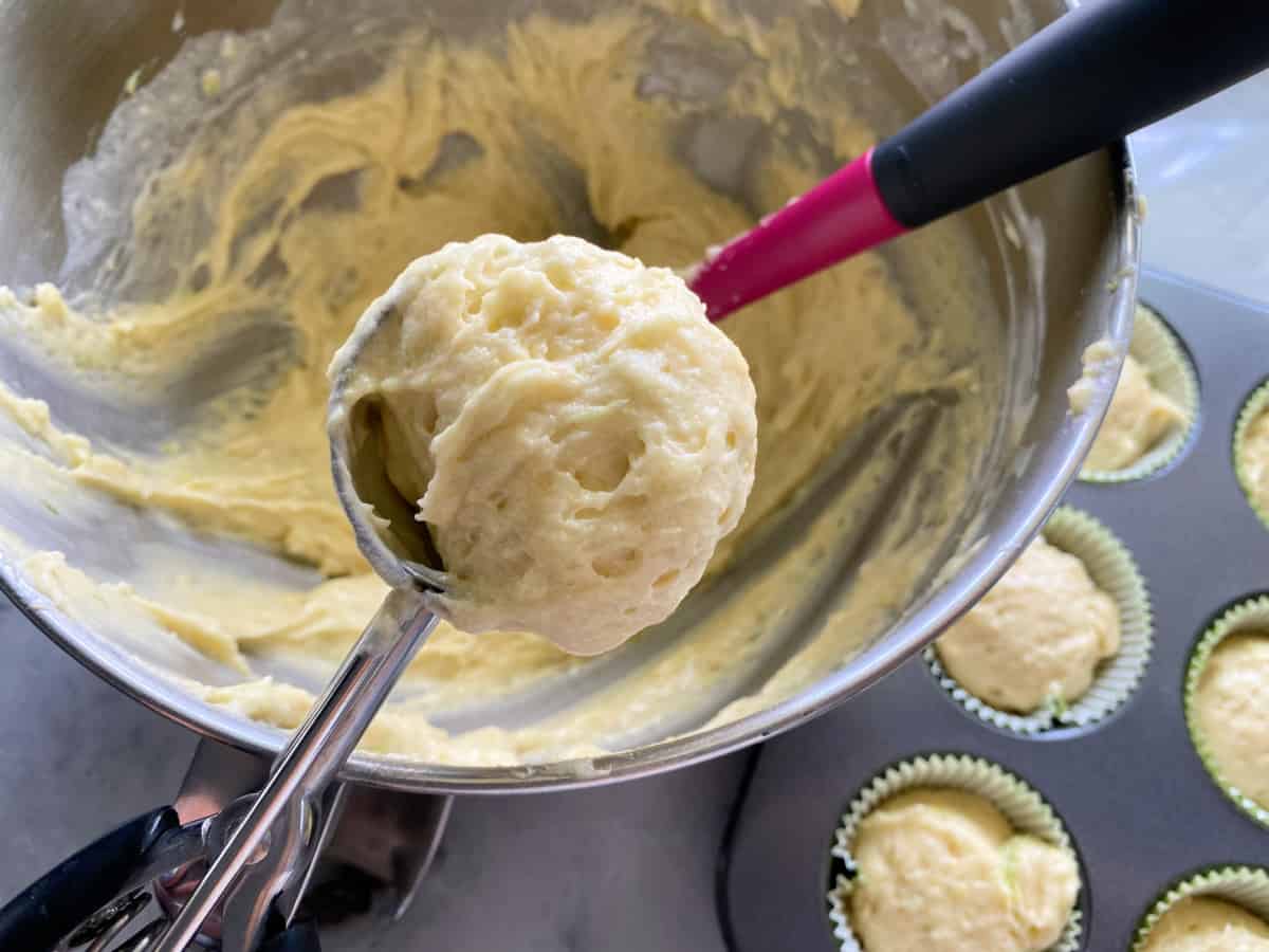 Cookie scoop with vanilla batter and raw cupcake batter in muffin tin below.