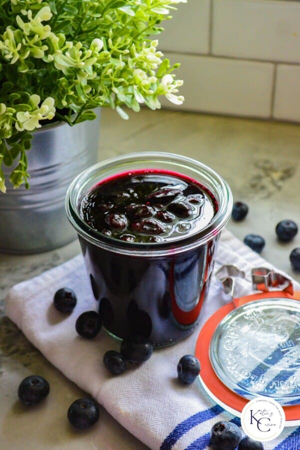 Glass jar filled with blueberry sauce resting on a white and blue cloth with logo on bottom right corner.