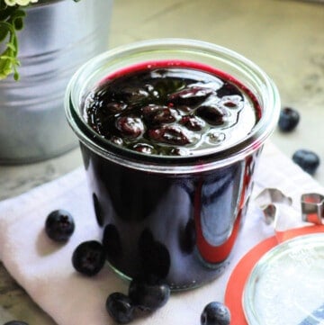 Glass jar filled with blueberry sauce with blueberries around it.