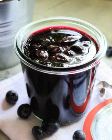 Glass jar filled with blueberry sauce with blueberries around it.