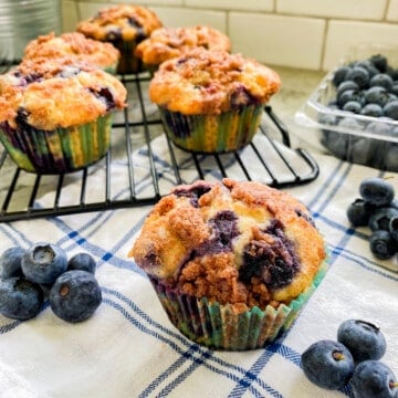 Blueberry muffin on white and blue cloth with other muffins on wire racks.