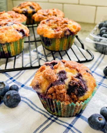 Blueberry muffin on white and blue cloth with other muffins on wire racks.