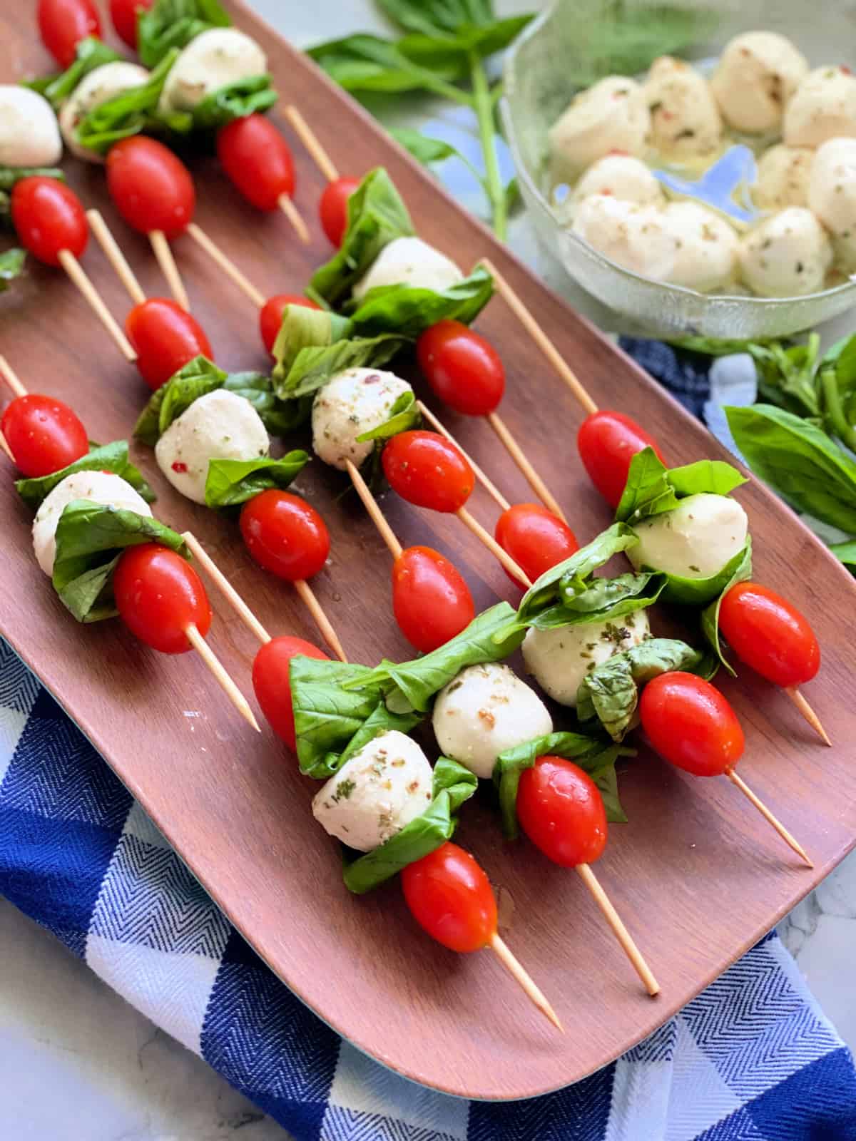 Caprese skewers on a platter on top of a blue and white checkered towel and mini mozzarella balls.