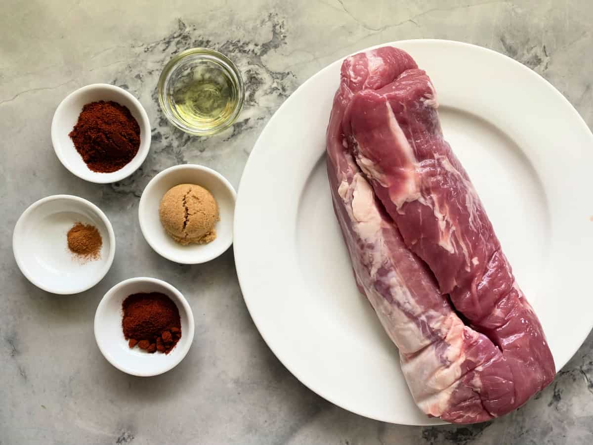 Ingrediens on counter; spices, oil, and pork tenderloin on white plate.