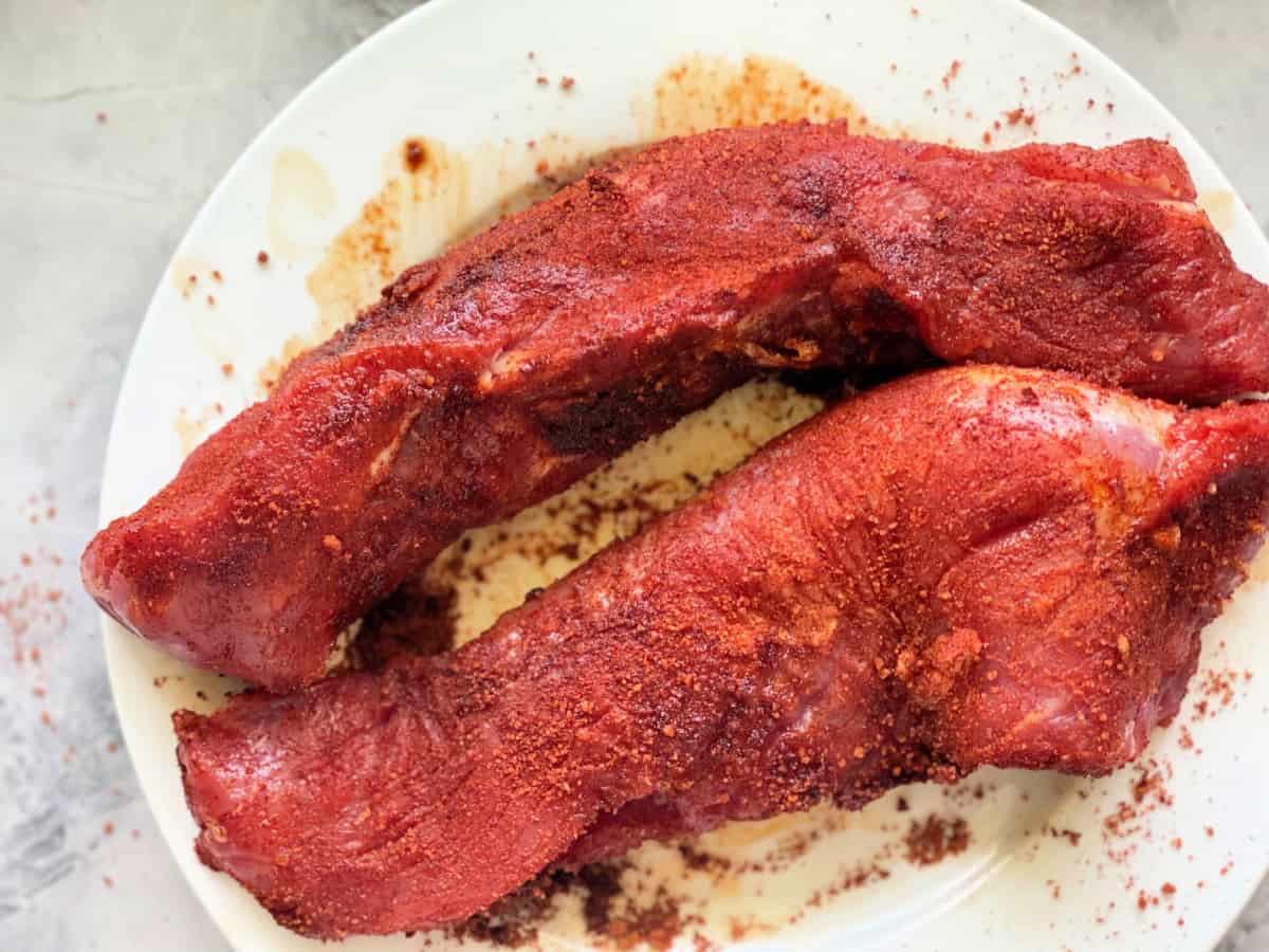 Two pieces of raw pork tenderloin with rub on it sitting on a white plate.