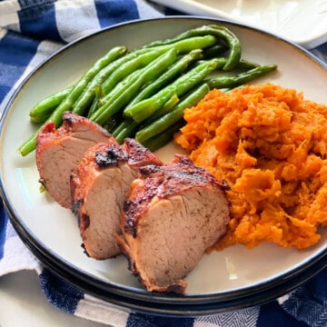 Sliced pork with green beans, and mashed sweet potato on a white and blue checkered cloth.