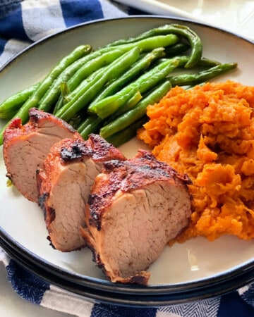 Sliced pork with green beans, and mashed sweet potato on a white and blue checkered cloth.