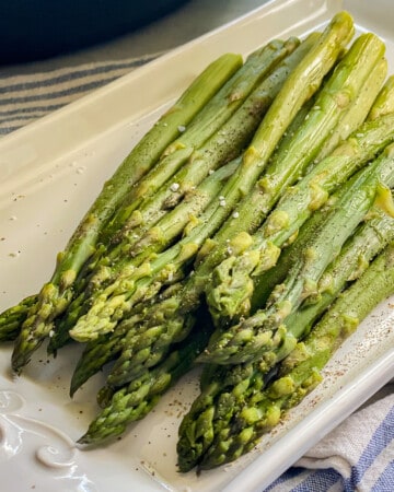 Cooked asparagus on a white platter topped with salt and pepper.