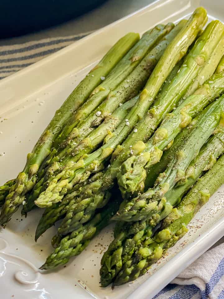 Cooked asparagus on a white platter topped with salt and pepper.