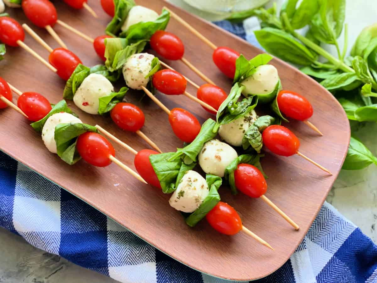 Brown platter with mozzarella, basil, and tomato skewers with blue and white checkered towel with basil on the bottom.
