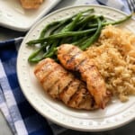 White plate with dots with 2 grilled chicken strips, rice, and green beans on a blue and white checkered cloth.