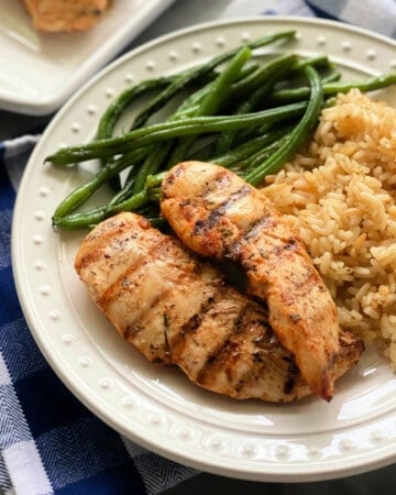 White plate with dots with 2 grilled chicken strips, rice, and green beans on a blue and white checkered cloth.