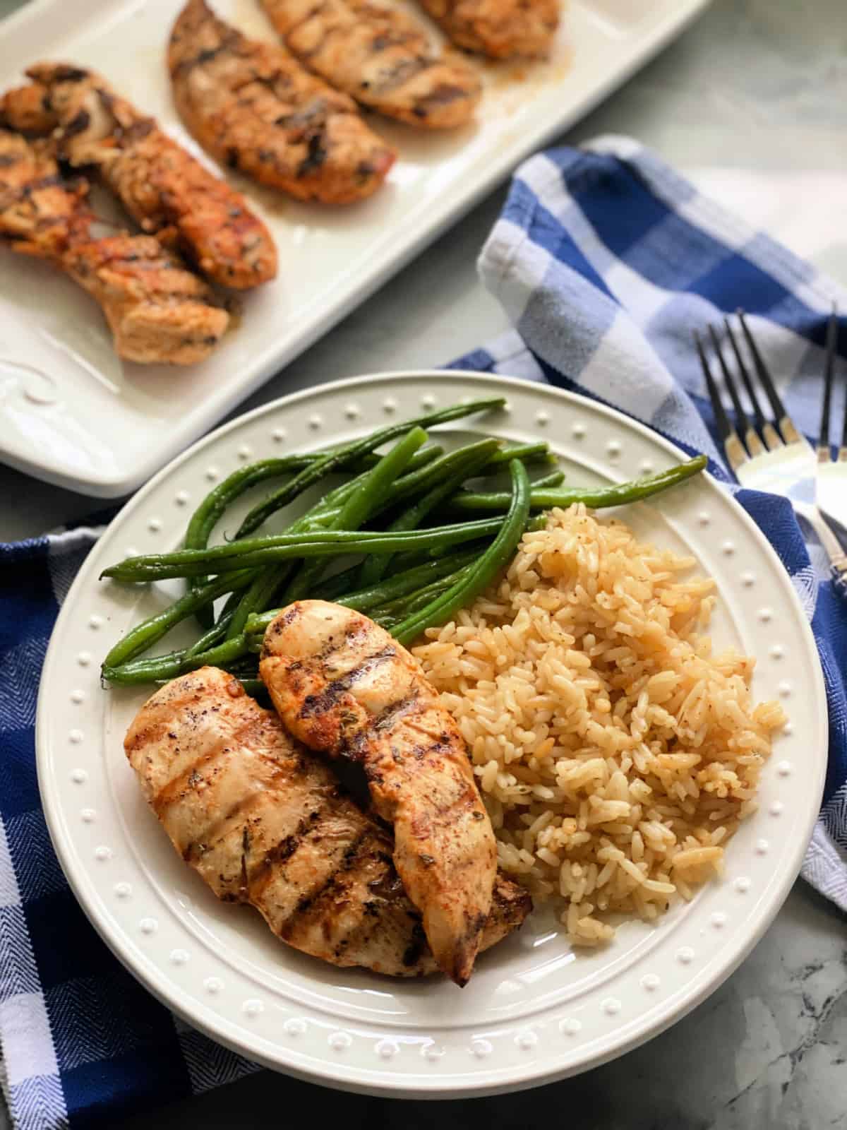 White round plate with 2 grilled chicken strips, rice, and green beans with a platter in the background with chicken strips.