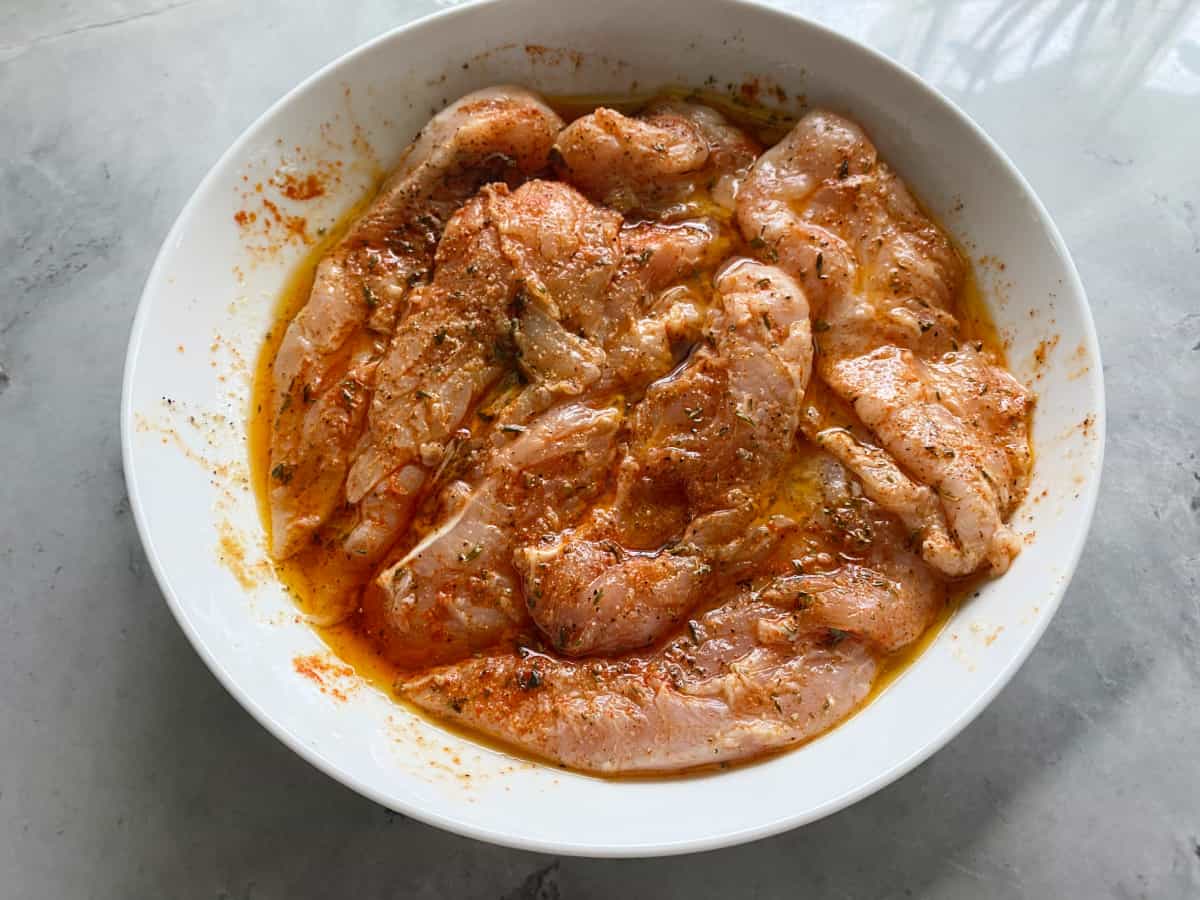 Round bowl filled with chicken strips in marinade on a marble countertop.