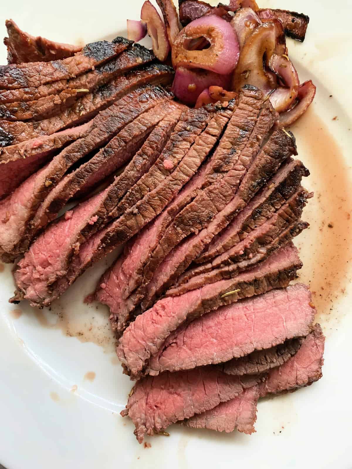 Thinly sliced rare steak with grilled onions next to it with steak juices on a white plate.