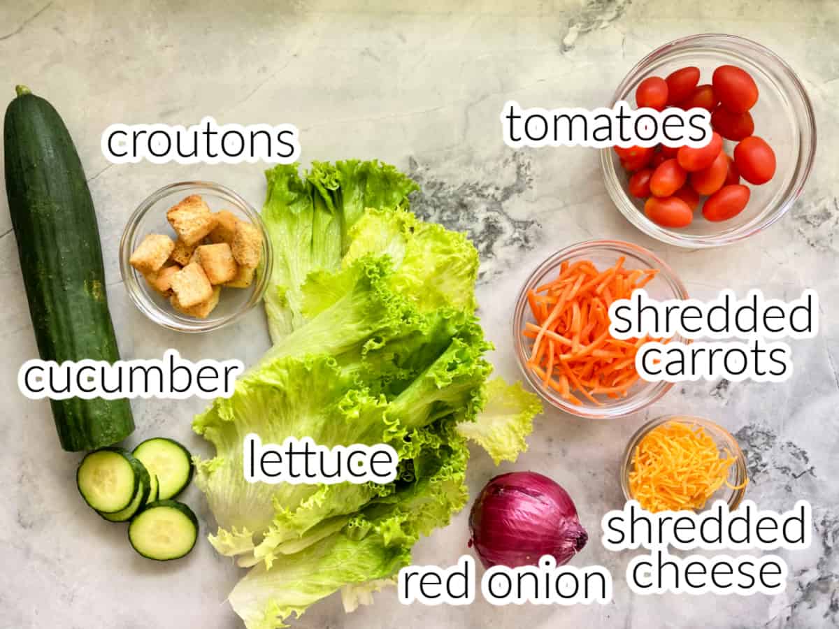 Ingredients on counter; cucumber, croutons, lettuce, red onion, shredded carrots, shredded cheese, and grape tomatoes.
