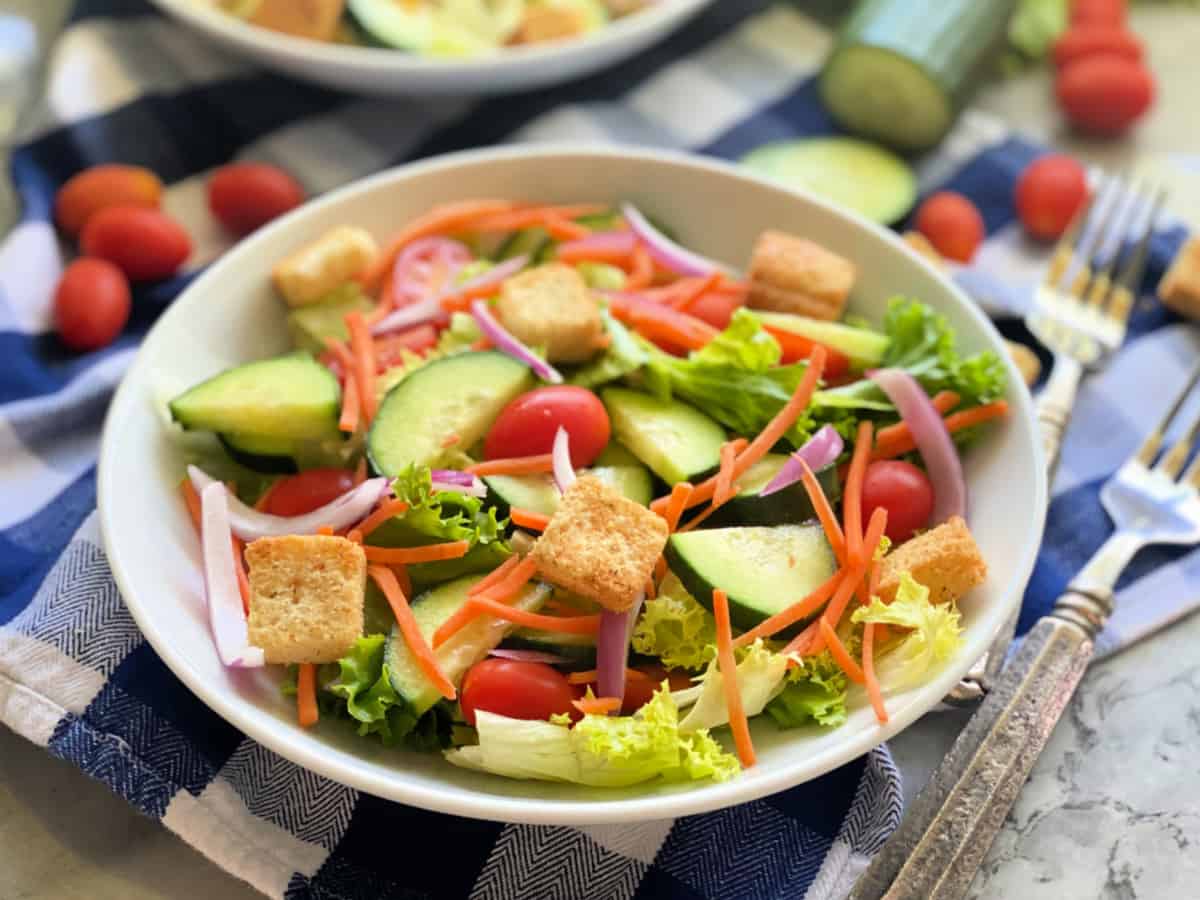 White large round bowl with salad on top of a blue and white checkered cloth with forks next to it.