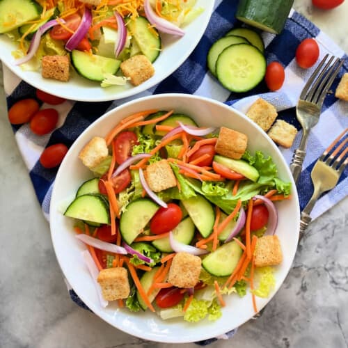 Two white bowls filled with salad and forks and veggies next to it.