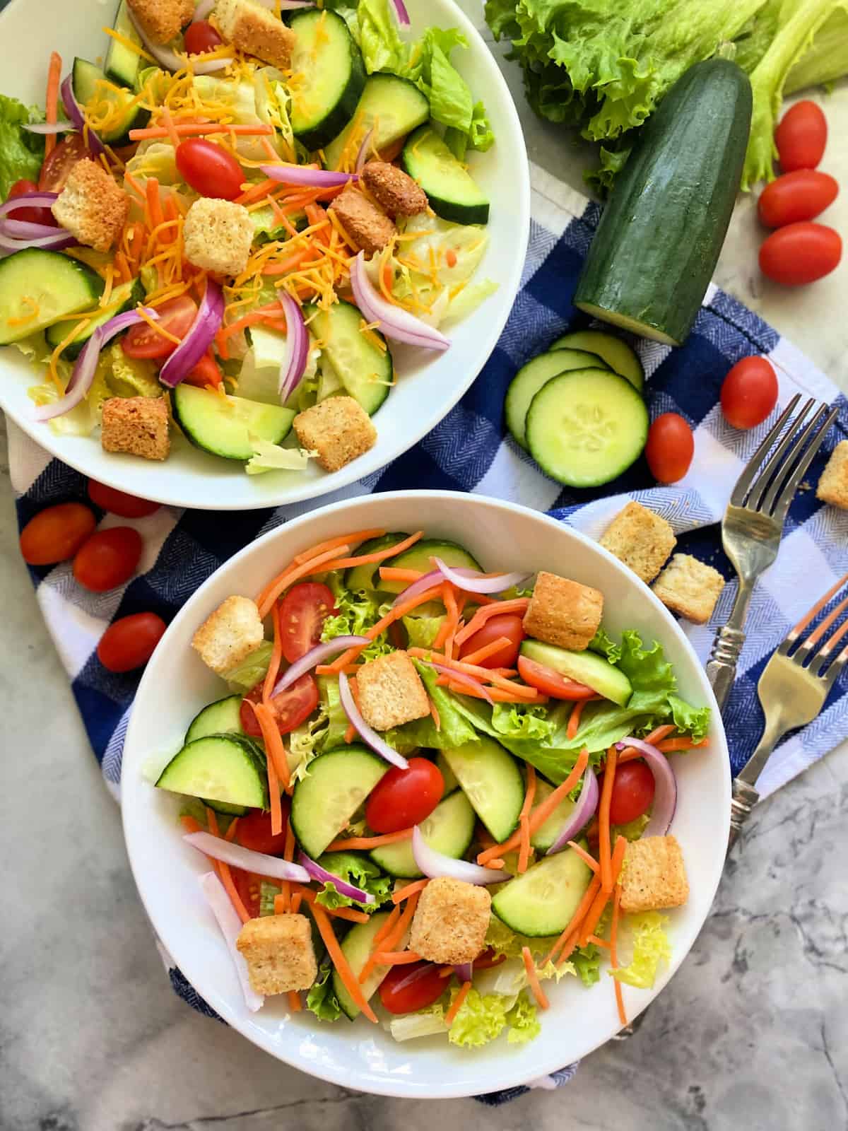 Two white bowls filled with lettuce, tomato, cucumber, onion, croutons, and cheese on top of a blue and white checkered cloth.