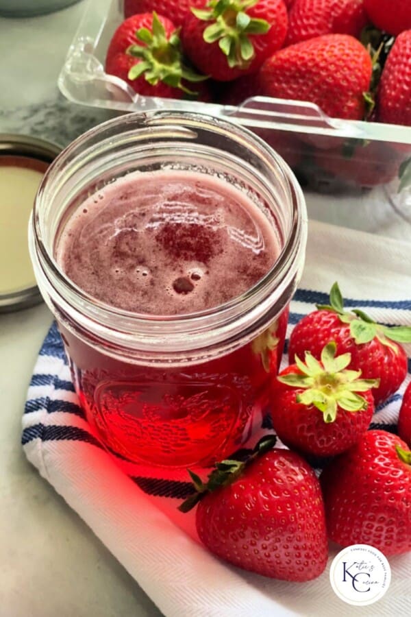 Pink liquid in a glass jar with strawberries around it and logo on bottom right corner.
