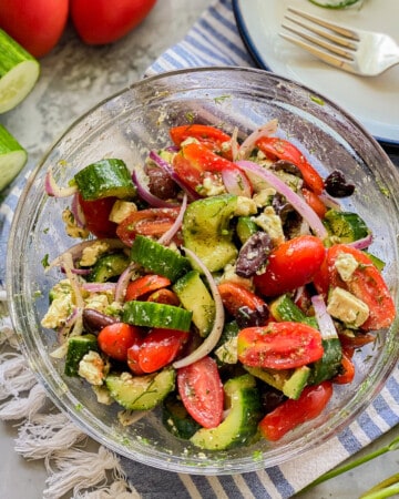 Glass bowl filled with cucumbers, tomatoes, olives, onions, and feta with cucumbers and tomatoes on the left corner.