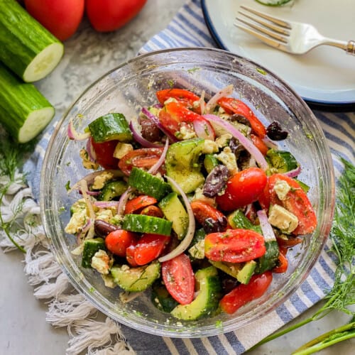 Glass bowl filled with cucumbers, tomatoes, olives, onions, and feta with cucumbers and tomatoes on the left corner.