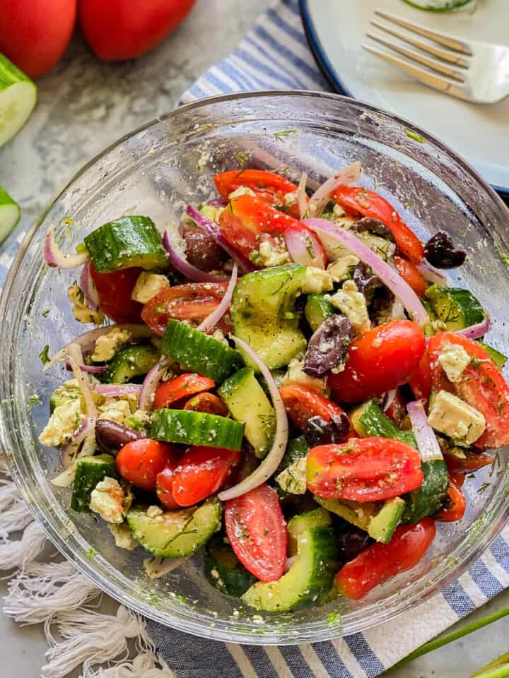 Glass bowl filled with cucumbers, tomatoes, olives, onions, and feta with cucumbers and tomatoes on the left corner.