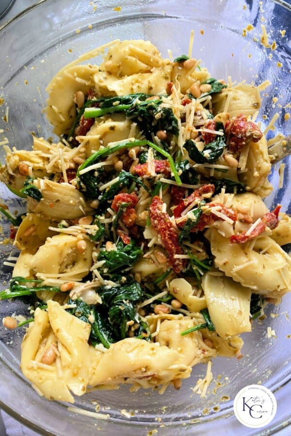 Glass bowl filled with tortellini salad with cheese, pine nuts, sun dried tomatoes, and spinach with logo on bottom right corner.