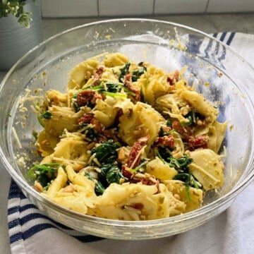 Glass bowl filled with tortellini salad resting on a white cloth with blue stripes.