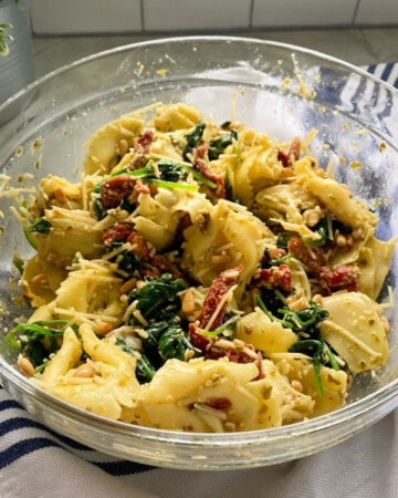 Glass bowl filled with tortellini salad resting on a white cloth with blue stripes.