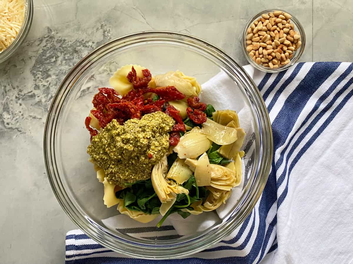 Glass bowl with pesto, sundried tomatoes, artichoke hearts, spinach and tortellini with blue and white cloth under bowl.