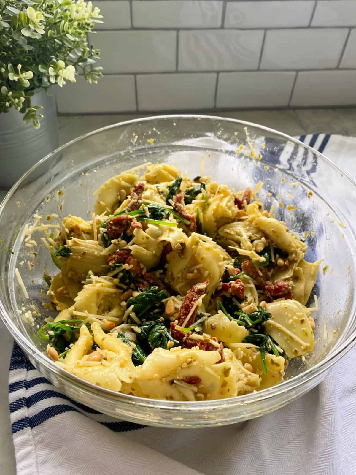 Glass bowl filled with a tortellini salad with vegetables on a marble countertop with a white subway tile back splash.