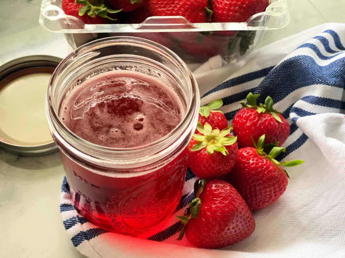 Glass mason jar filled with pink liquid resting on a white and blue striped cloth with fresh berries next to it.