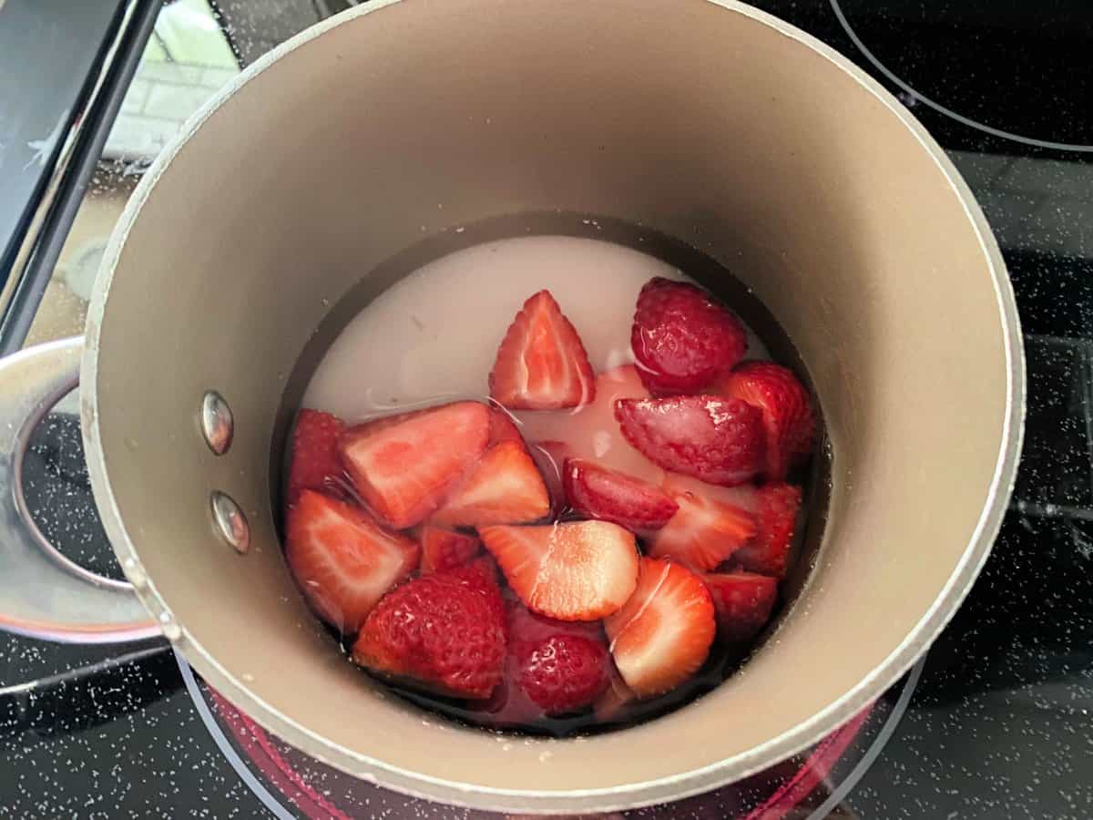 Chopped strawberries in a pot with water, and sugar on a stovetop.