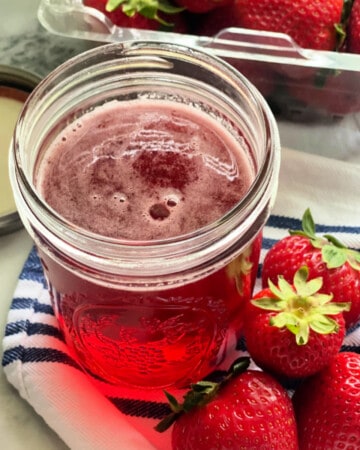 Glass mason jar sitting on a white cloth filled with pink liquid and strawberries next to it.