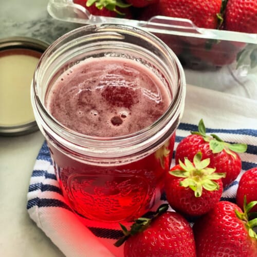 Glass mason jar sitting on a white cloth filled with pink liquid and strawberries next to it.
