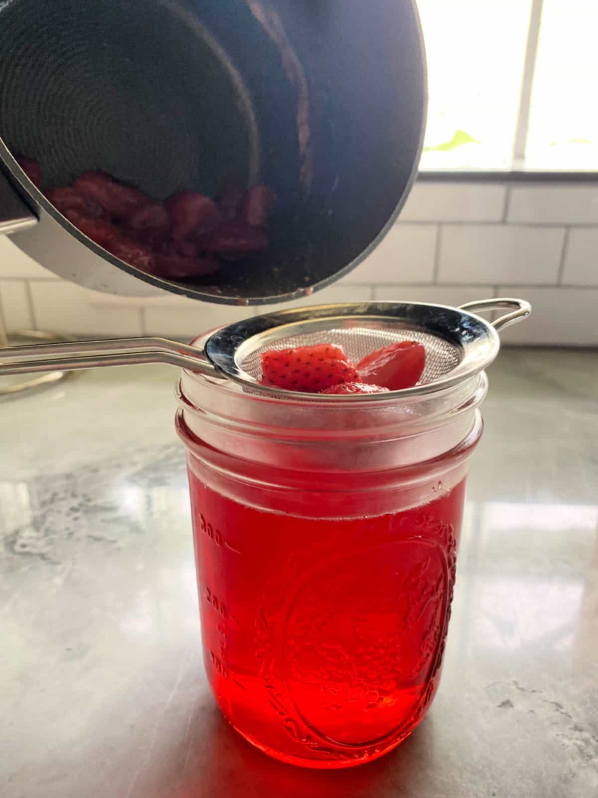 B lack pot pouring liquid and strawberries over a strainer on a glass mason jar.