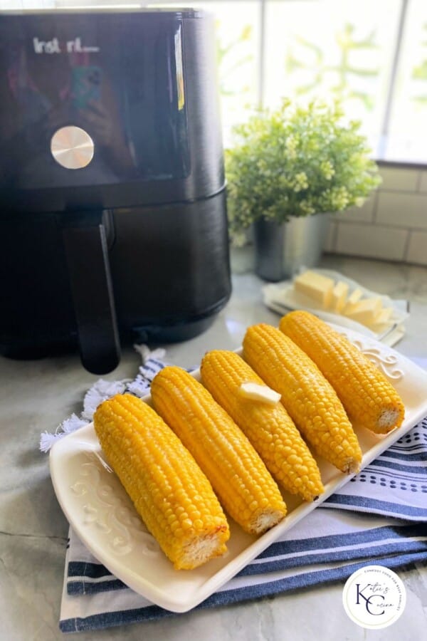 5 corn on the cobs on a long white dish with a pad of butter on top with a black air fryer and slices of butter on the side